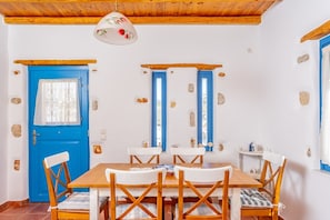 A dining area with a wooden table and white chairs, a blue door, and a stone wall. - Villa Ioanna for 10 - Allparos Villas (Paros)
