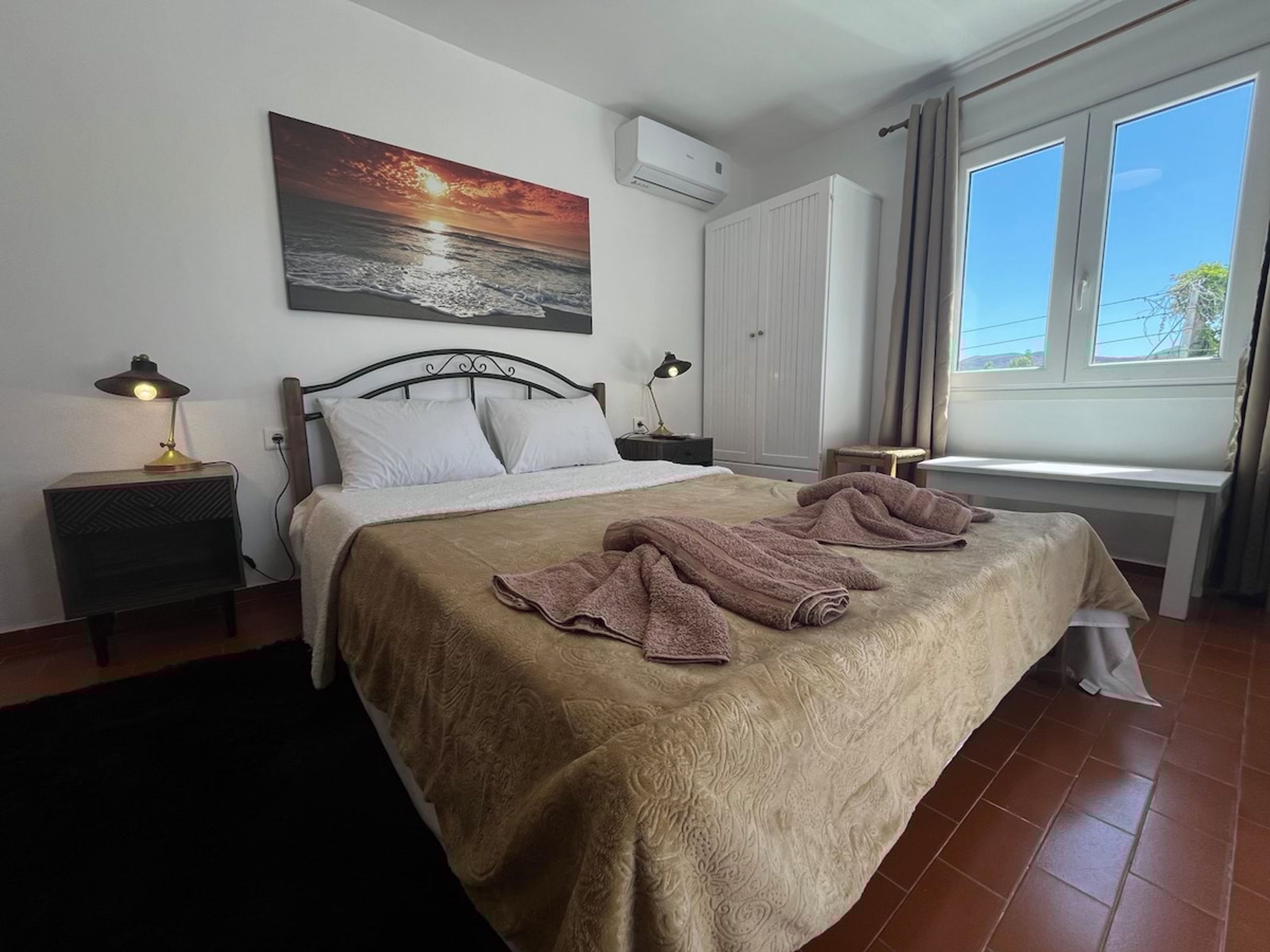 A neatly made bed with a patterned bedspread, two bedside lamps, a wall-mounted air conditioner, a window with curtains, and a painting of a sunset over the sea.