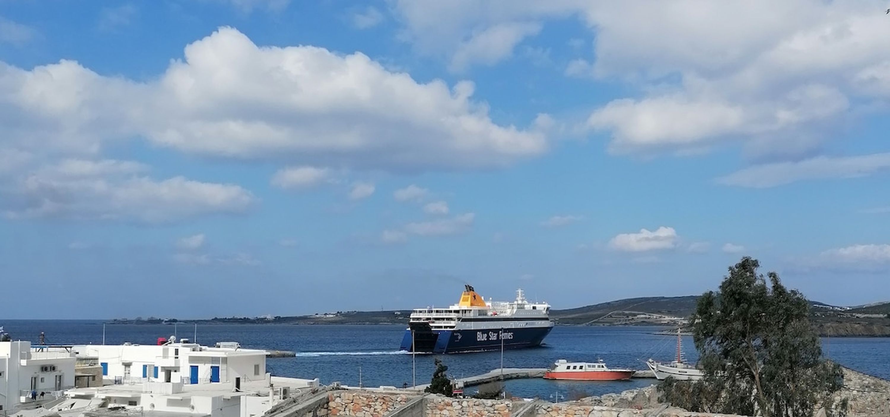 A coastal scene with a ferry named "Blue Star Ferries" docked at a port, surrounded by buildings and a clear sky.