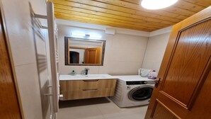 A compact bathroom with a wooden ceiling, a built-in washing machine, a sink with a mirror, and a wooden door. - Cozy Apartment with mountain view (Bad Kleinkirchheim)