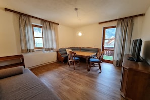A cozy room with a wooden floor, a sofa, a dining table with chairs, a bench, and a television on a wooden cabinet.