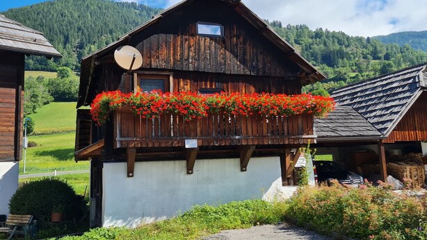 A wooden chalet with a balcony adorned with red flowers, a satellite dish, and a view of a green valley and mountains. - Apartment at the ski slope, Bad Kleinkirchheim (Bad Kleinkirchheim)