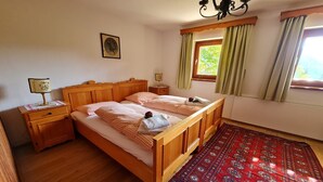 Een houten bed met een geruit beddegoed, een nachtkastje met een lamp, een raam met gordijnen, en een kamerbreed tapijt met dessin.