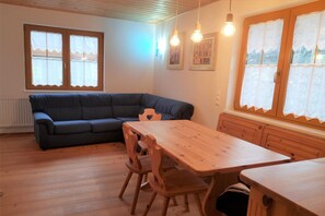 A cozy room with a wooden floor, a dark blue sofa, a wooden dining table with chairs, and a window with lace curtains.