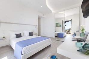 A modern hotel room with a large bed, a desk with a laptop, and a view of the sea through an open door.