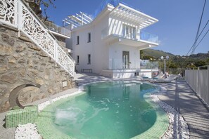 A modern building with a pool, stone wall, and a balcony with a view of the hills.