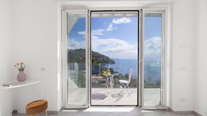 Ein Zimmer mit einer Schiebeglastür, die auf einen Balkon mit Blick auf das Meer und eine hügelige Landschaft führt.