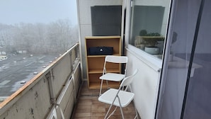 Een balkon met betegelde vloer, een witte klapstoel, een klein houten tafeltje en een raam met uitzicht op een parkeerplaats.