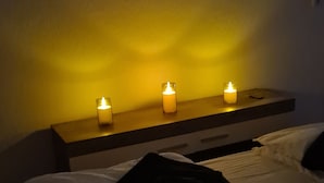 Een bed met witte lakens en een zwarte deken, een nachtkastje met drie brandende kaarsen, en een wand met een structuur afwerking.