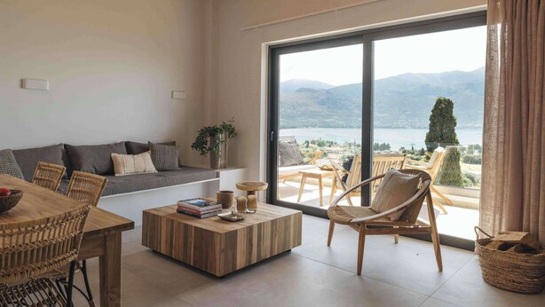Ein geräumiges Wohnzimmer mit großem Fenster, das Blick auf Berge und einen See bietet. Es gibt einen Holztisch, eine Couch mit Kissen sowie Korbstühle.