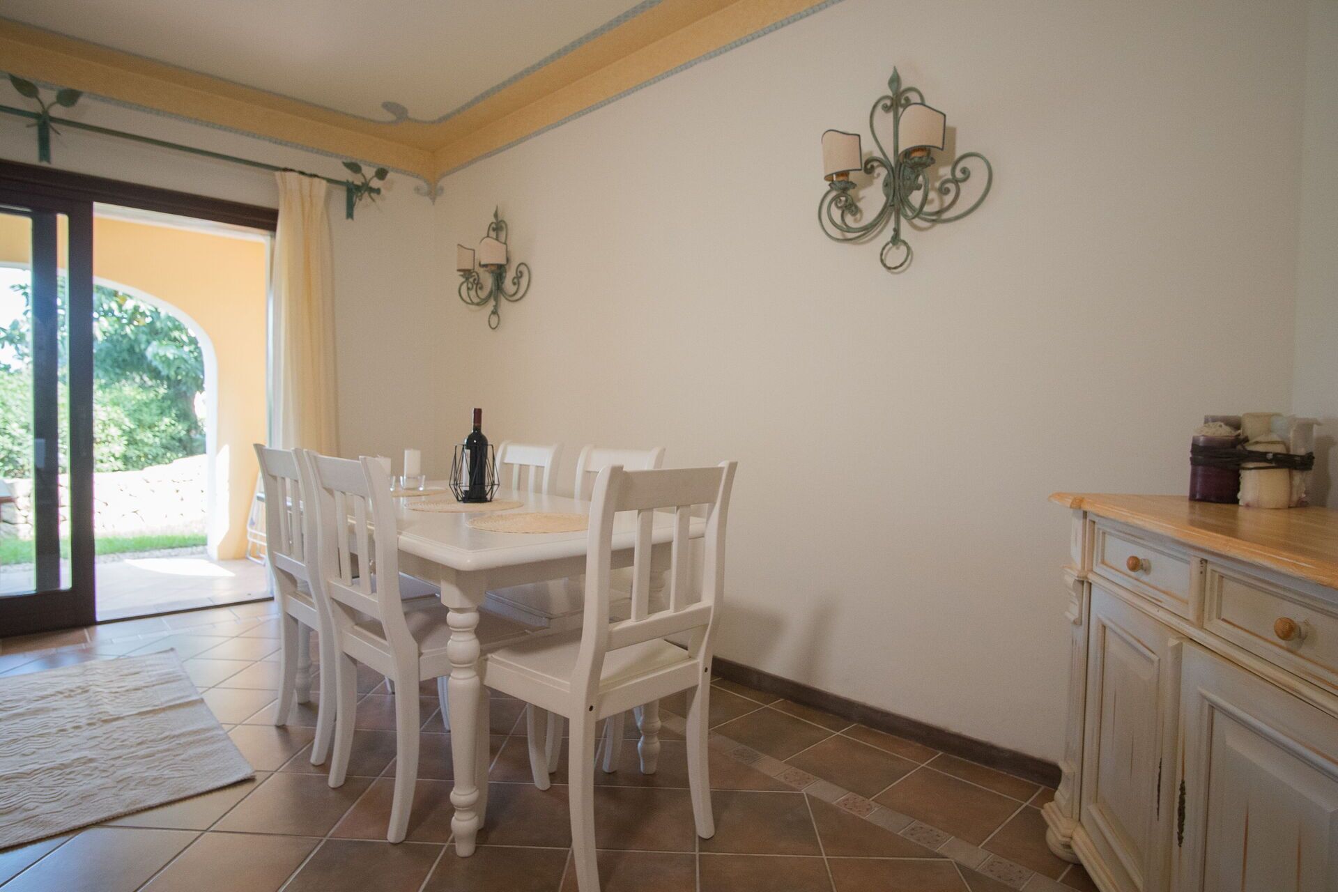 Un coin repas avec une table et des chaises blanches, une bouteille de vin et un meuble avec des tiroirs.