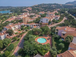 An aerial view of a coastal residential area with terracotta-roofed houses, a central swimming pool, and a road leading to the sea. - Beautiful apartment for 4 people with pool, WIFI, A/C and TV (Porto Cervo)