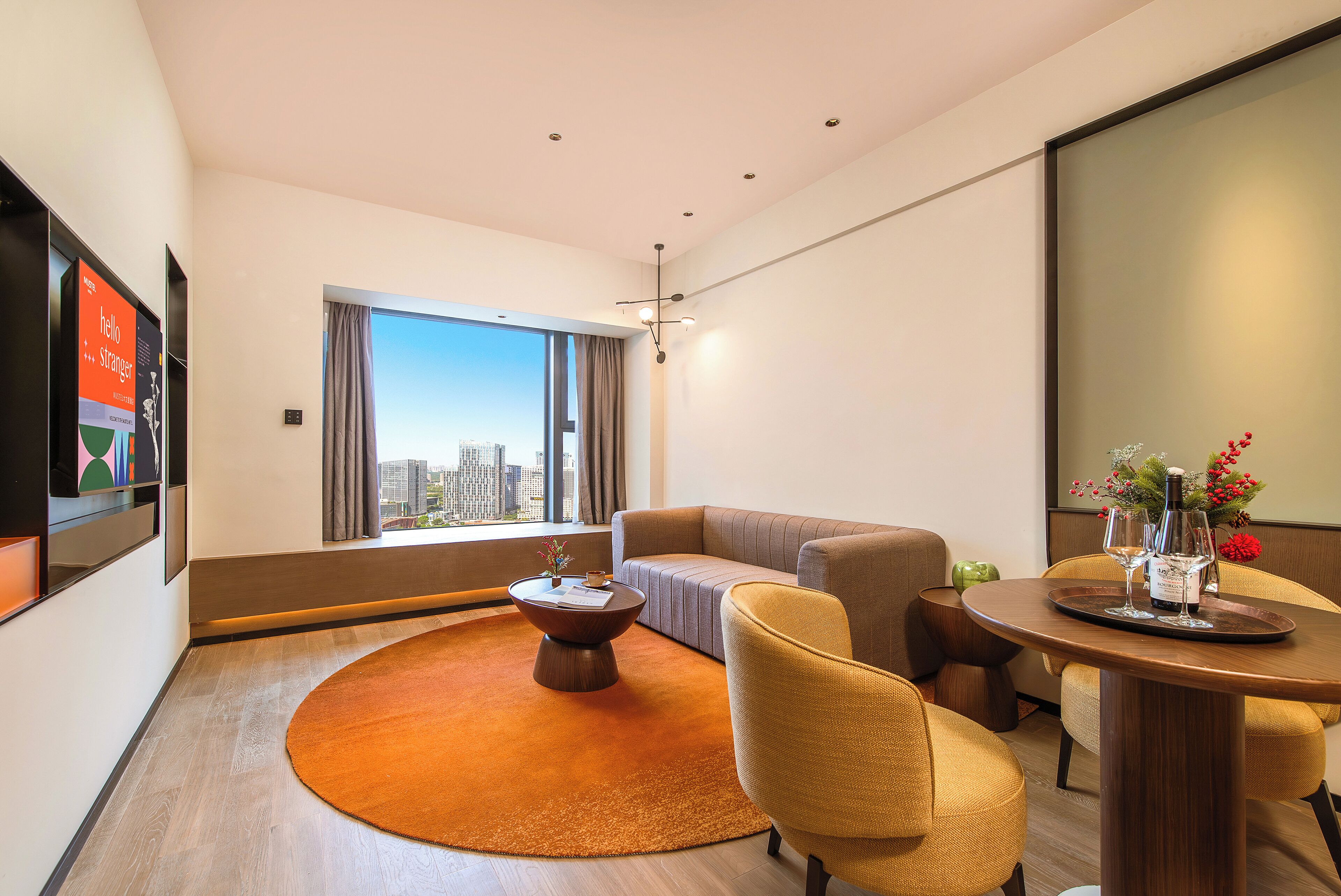 A modern hotel room with a large window, a sofa, a round coffee table, and a dining table with a tray.