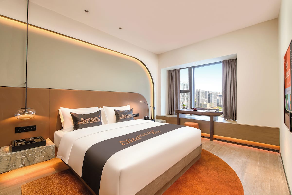 A modern hotel room with a large bed, a city view, a desk, and a window with curtains.