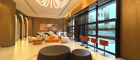 Eine moderne Lobby mit einem Marmor-Rezeptionstresen, Sitzbereichen mit orangefarbenen und schwarzen Hockerpoufs sowie großen Fenstern mit Blick ins Freie.