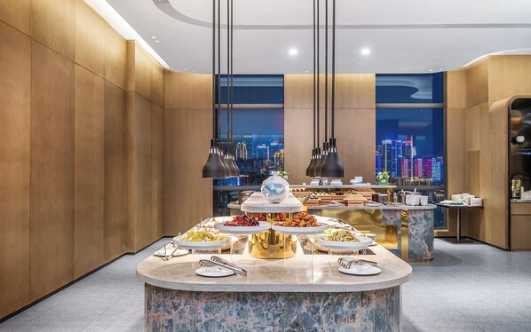 A modern dining area with a marble counter displaying various dishes, a cityscape view through large windows, and pendant lighting. - MUSTEL  ARTE  HOTEL  ZHENLIN  CHENGDU (Chengdu)