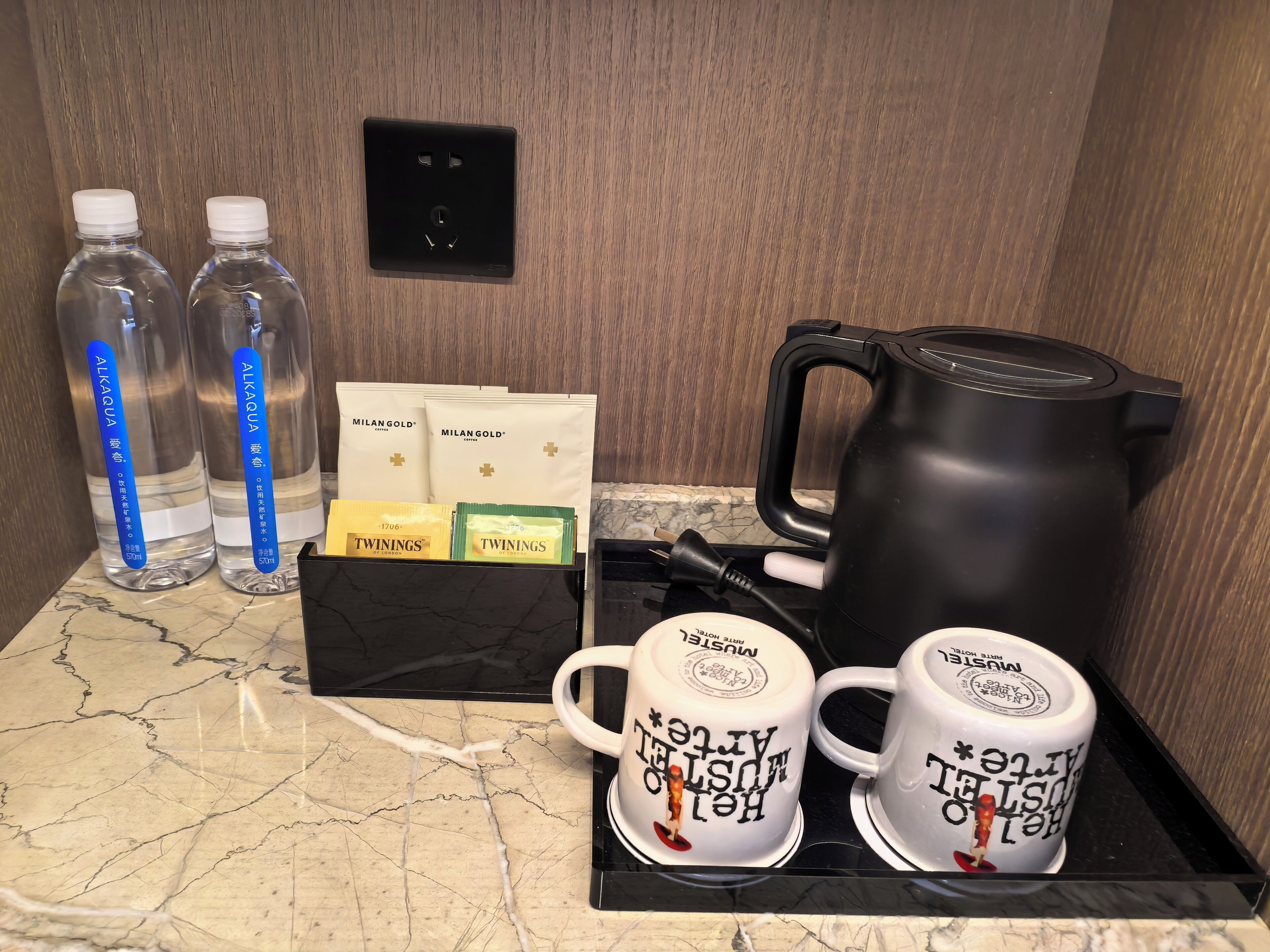 A hotel room's minibar with bottled water, coffee packets, and a black electric kettle on a marble countertop.