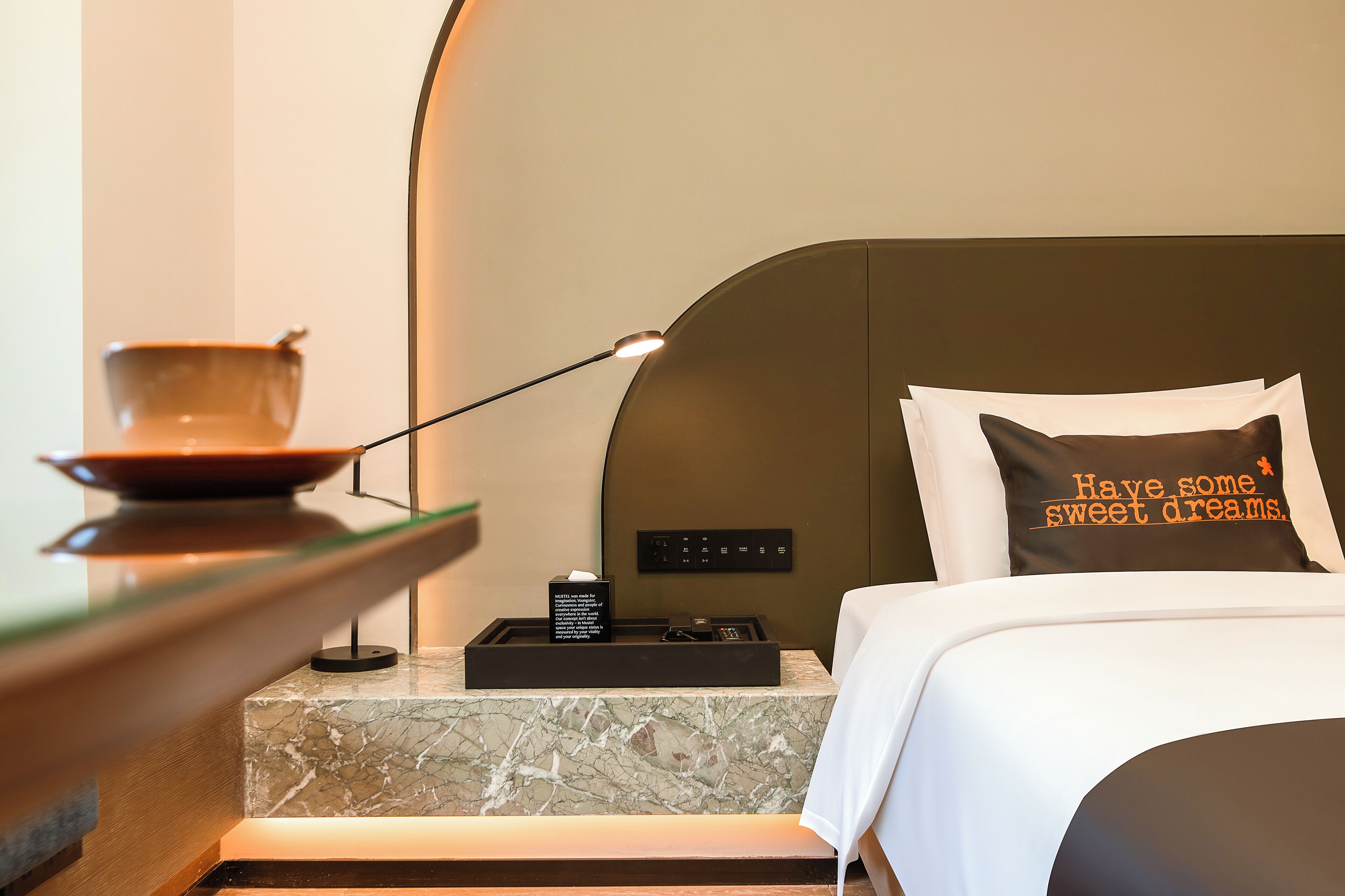 A hotel room with a marble bedside table, a bowl, a remote control, and a pillow with a motivational quote.