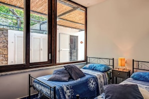 A bedroom with two beds, a window with shutters, a nightstand with a lamp, and a view of a stone wall and door outside.