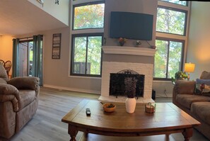 A spacious living room with a fireplace, large windows, and a flat-screen TV mounted above it. - Welcome To "Snowed Inn” - Walk On And Off The Slopes (McGaheysville)