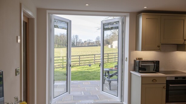 A kitchen with a microwave and oven, a dining area with a table and chairs, and a view of a grassy field through open doors. - The Dog Kennels, Leyburn (Leyburn)