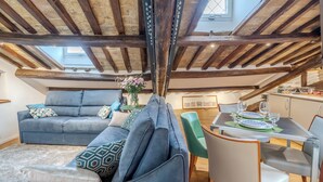 An attic living space with exposed wooden beams, a grey sofa set, a dining table with chairs, and a cityscape painting. - Elegant historic apartment, Dimora Capo di Ferro (Roma)