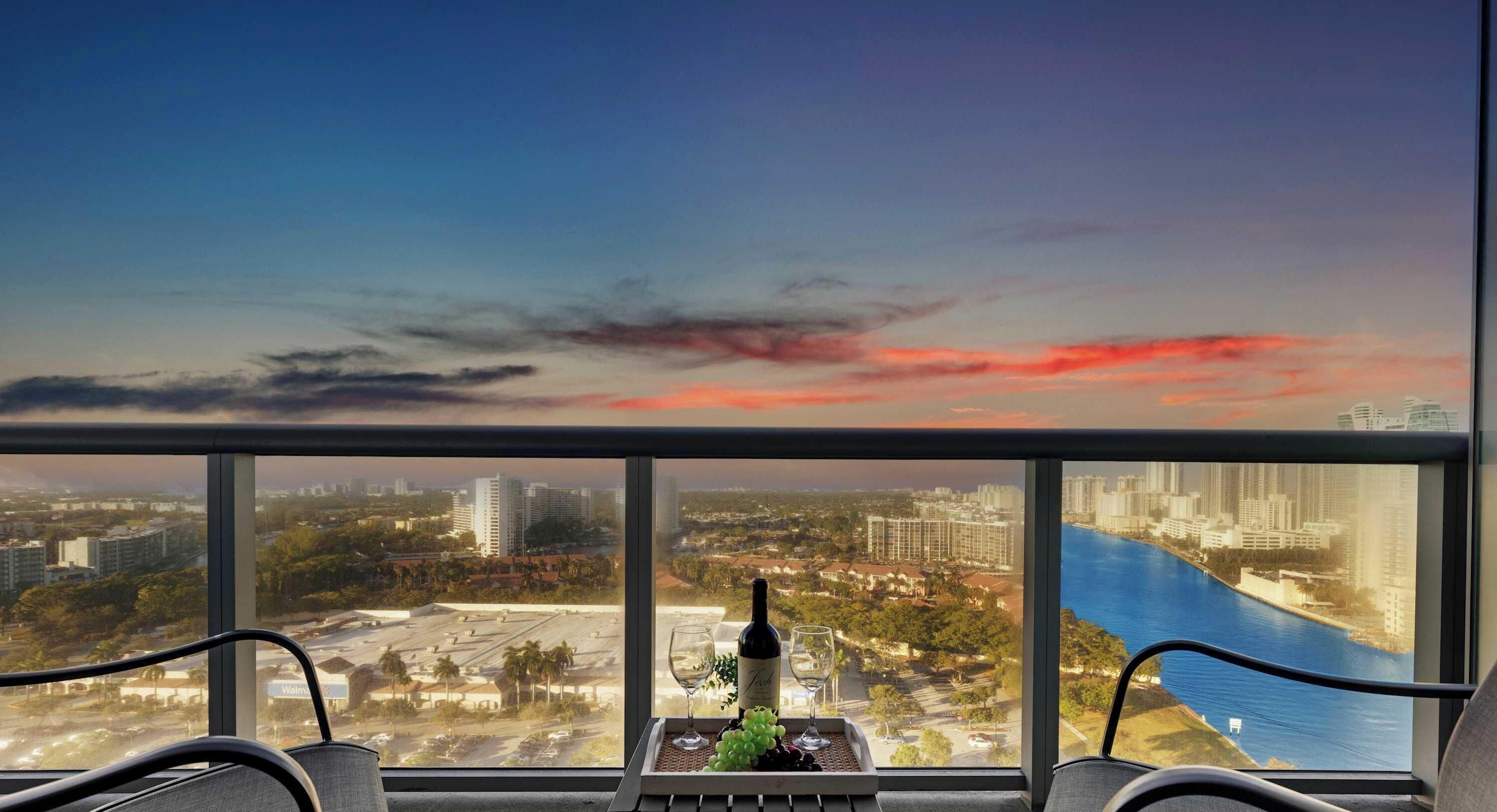 A cityscape view from a high-rise building with a balcony, a table with a bottle of wine and glasses, and a river in the foreground.