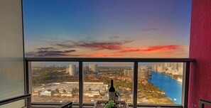 A view from a high-rise window overlooking a cityscape with a river, a sunset, and a bottle of wine on a table.