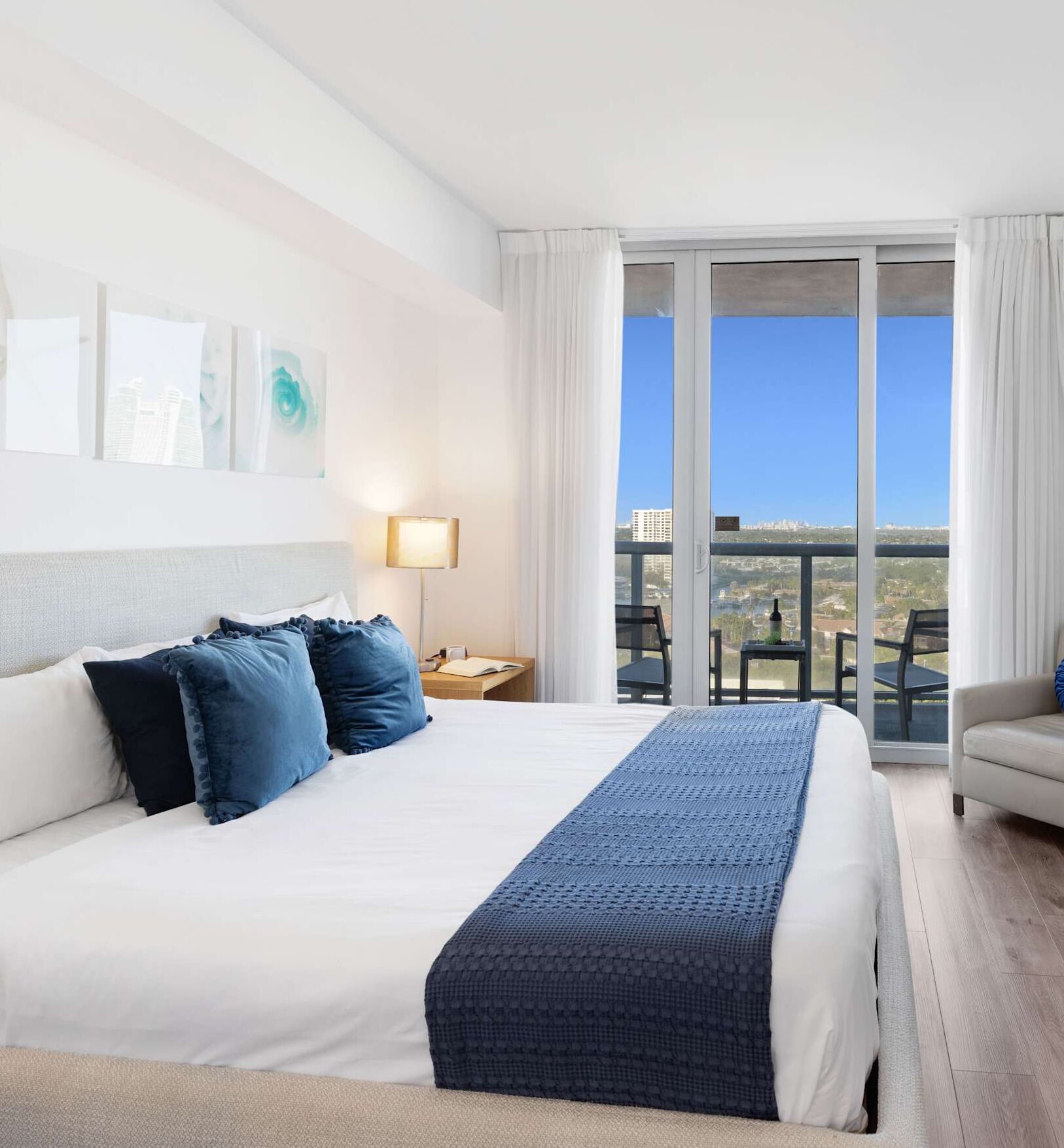 A hotel room with a large bed, a nightstand, a lamp, a view of the city through large windows, and a balcony with outdoor furniture.