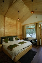Quarto com painéis de madeira, uma cama grande, duas cadeiras, uma mesinha e uma janela com cortinas.
