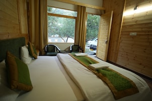 Quarto com uma cama grande, duas cadeiras, janela com vista para árvores e carros estacionados, e paredes de madeira.