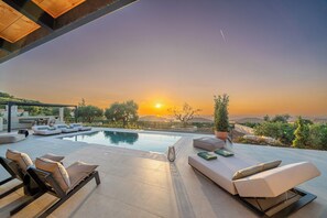 A modern outdoor pool area with lounge chairs and a dining setup, overlooking a scenic landscape during sunset.