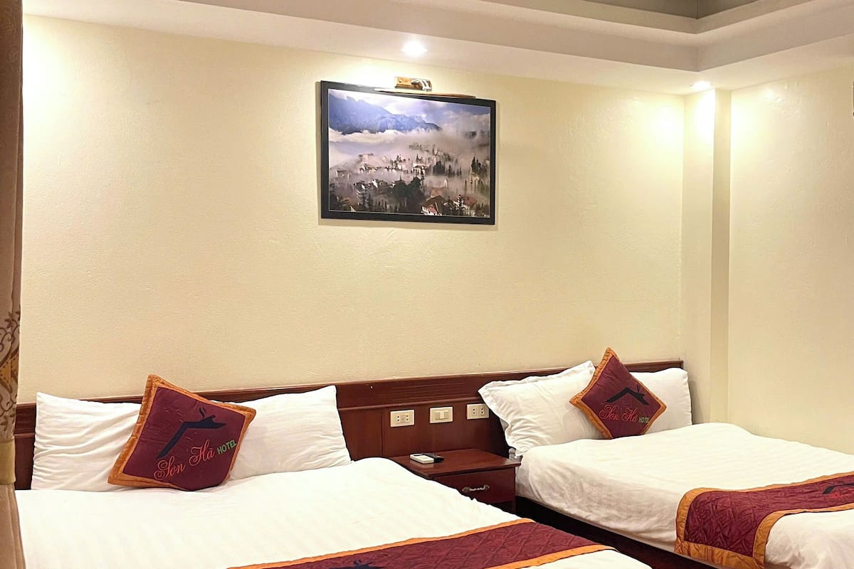 A hotel room with two beds, each with white bedding and a red and gold bed runner. There is a painting of a mountain landscape on the wall above the beds.