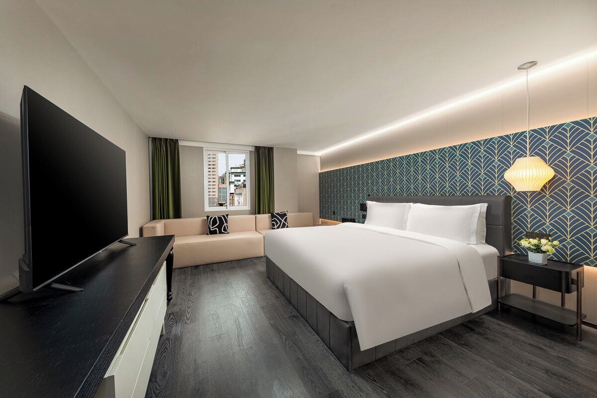 A modern hotel room with a large bed, a flat-screen TV, a seating area, and a city view.