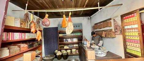 A rustic shop with hanging meats, shelves of packaged goods, and a counter with a person preparing food.