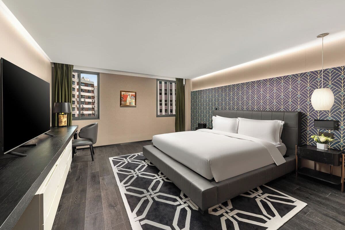 A modern hotel room with a large bed, a desk with a flat-screen TV, a chair, and a patterned rug.