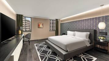 A modern hotel room with a large bed, a desk with a flat-screen TV, a chair, and a patterned rug.