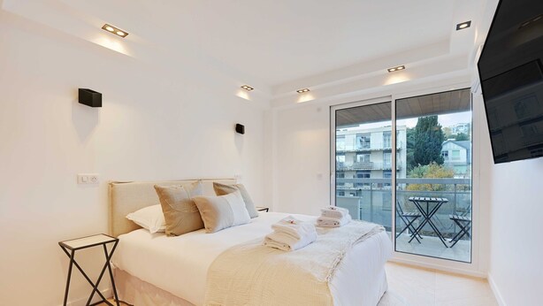 A modern bedroom with a large bed, a small side table, and a balcony with a table and chairs. - Charming Apt With AC & Balcony (Suresnes)