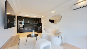 A modern living room with a sofa, ottoman, and coffee table, a kitchen area with dark cabinetry and appliances, and a large mirror on the wall. - Charming Apt With AC & Balcony (Suresnes)
