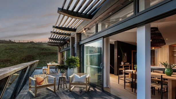 Un patio moderno al aire libre con mobiliario de madera, una puerta de cristal que da a un comedor interior y vistas a una colina cubierta de césped.