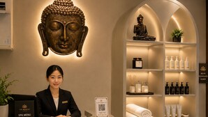 Un comptoir d’accueil avec un réceptionniste, une sculpture représentant une tête de Bouddha, un menu de soins de spa, ainsi que des étagères contenant des articles de toilette et des serviettes.