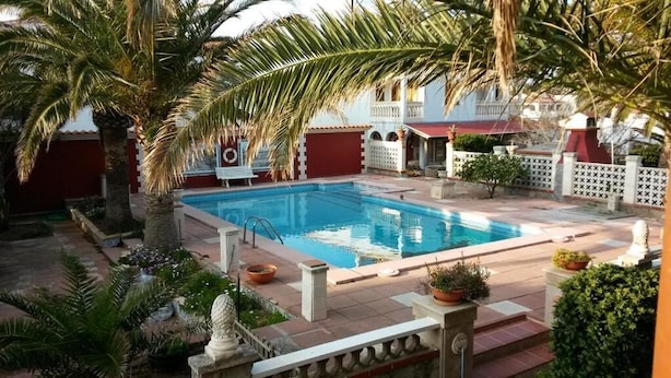 A swimming pool surrounded by palm trees and a red building with a balcony. - Las Belisas (Mercadal)