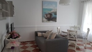 A living room with a grey sofa, a dining table with chairs, a painting of a boat on the wall, and a vase of flowers on a side table. - Las Belisas (Mercadal)