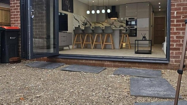 A modern kitchen with a bar area, large windows, and a visible outdoor area with gravel. - Chester - 4 Bed House Free Parking (Chester)