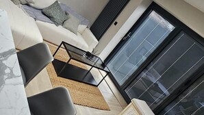A modern living room with a sofa, chairs, a coffee table, and a TV. There is a large window with blinds and a skylight. - Chester - 4 Bed House Free Parking (Chester)