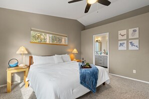 Une chambre à coucher comprenant un lit, deux tables de chevet, un ventilateur de plafond, une fenêtre avec vue, une salle de bain avec un miroir et des œuvres d’art accrochées au mur.