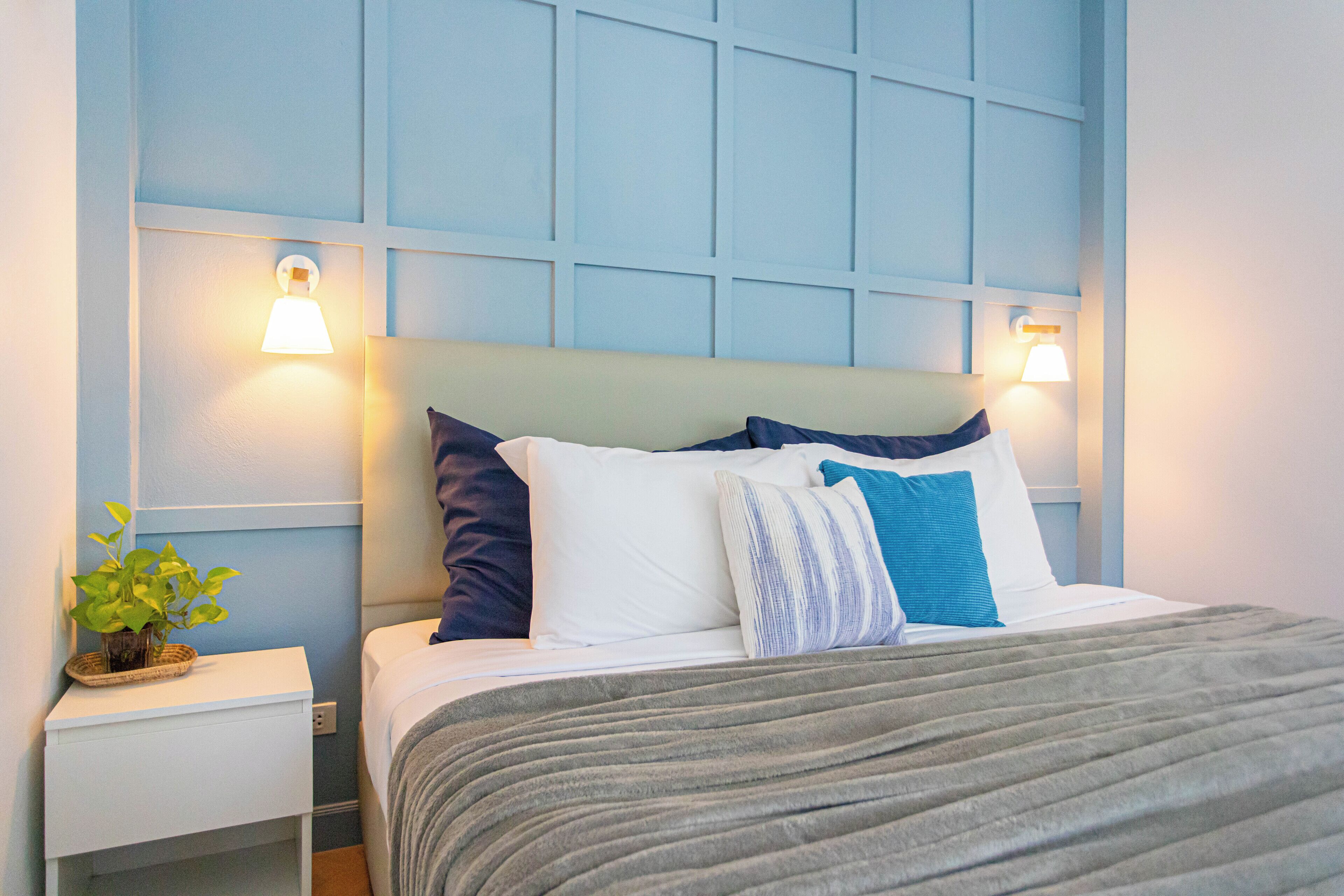A neatly made bed with a grey blanket and multiple pillows, a small bedside table with a plant, and wall-mounted lights.