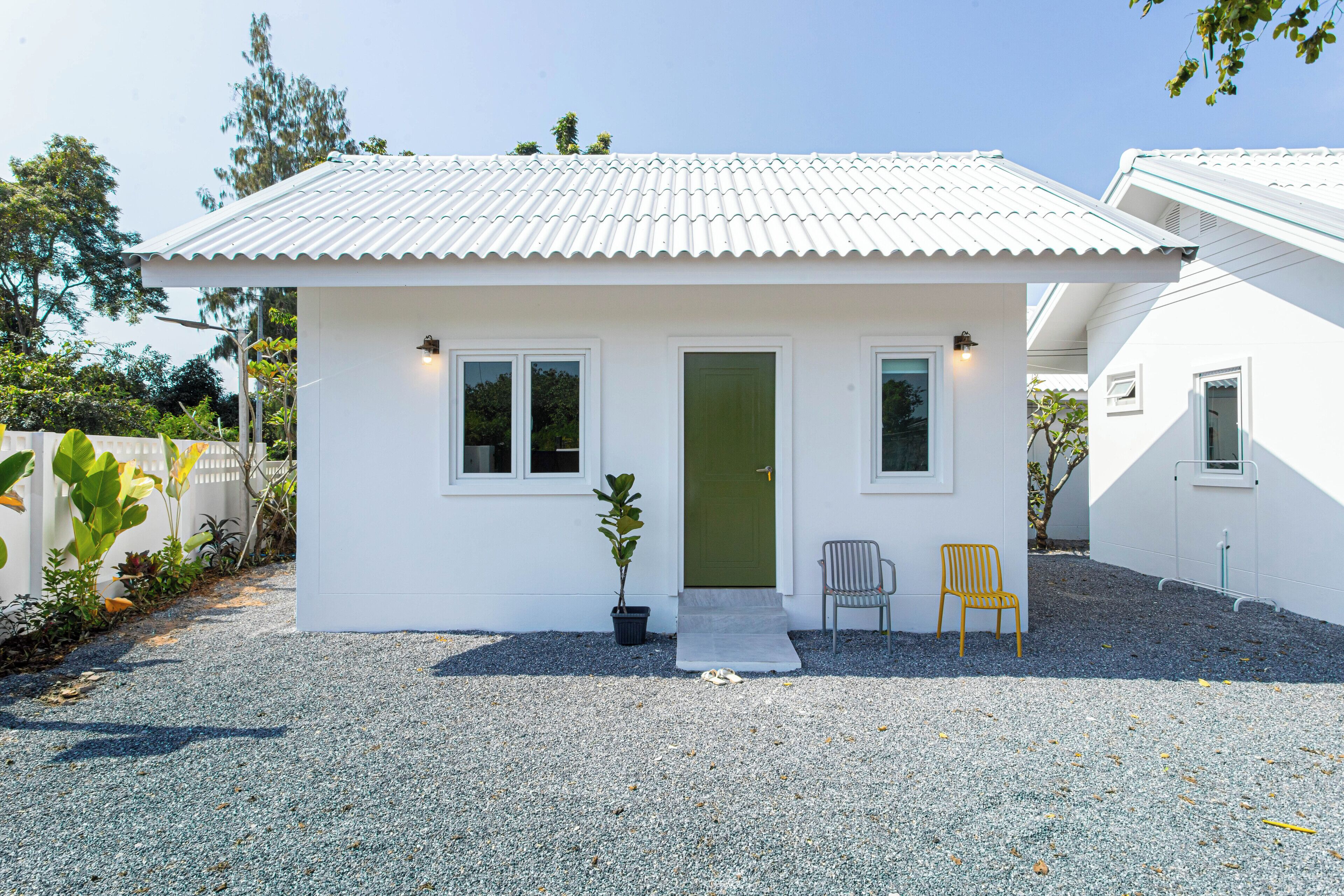 Een laag, enkel bouwlaag huis met een witte gevel, een groene deur en een grindpad. Er staan twee stoelen buiten en een bloempot bij de ingang.