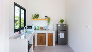 A compact kitchen with a microwave, toaster, and refrigerator, a window with blinds, and a small shelf with plants and kitchen items. - Vawa Guesthouse (Kanchanaburi)
