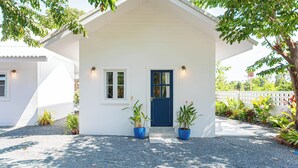 A small white house with a blue door and windows, surrounded by gravel and potted plants. - Vawa Guesthouse (Kanchanaburi)